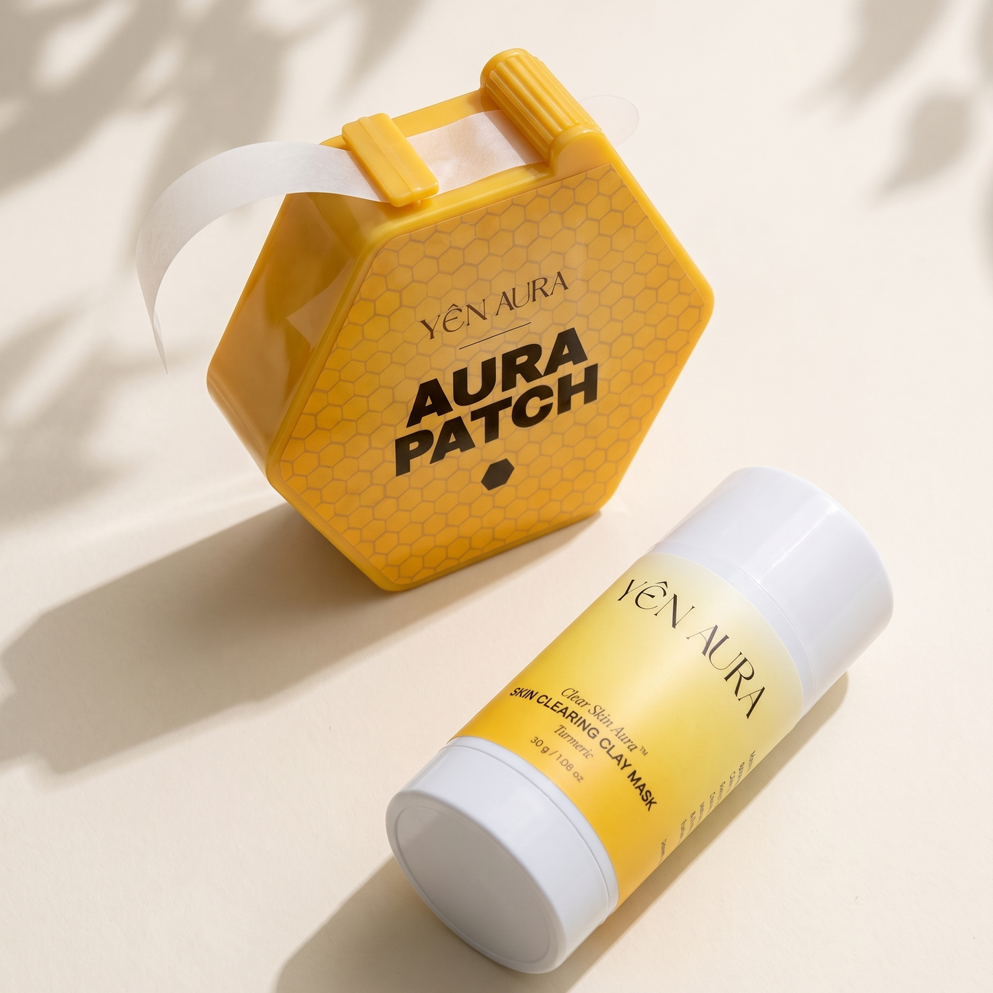 Yen Aura Patch sheet mask set, featuring a yellow hexagonal patch box with a white strip unrolled from it, and a matching yellow and white stick of facial sheet mask, styled on a warm beige background with leaf shadows.